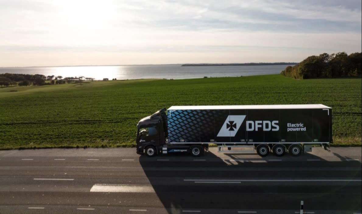 A blue truck, displaying the DFDS logo and the text "Electric powered" on the sside, drives past a green terrain with a lake on the background DFDS electric powered truck, cropped hero