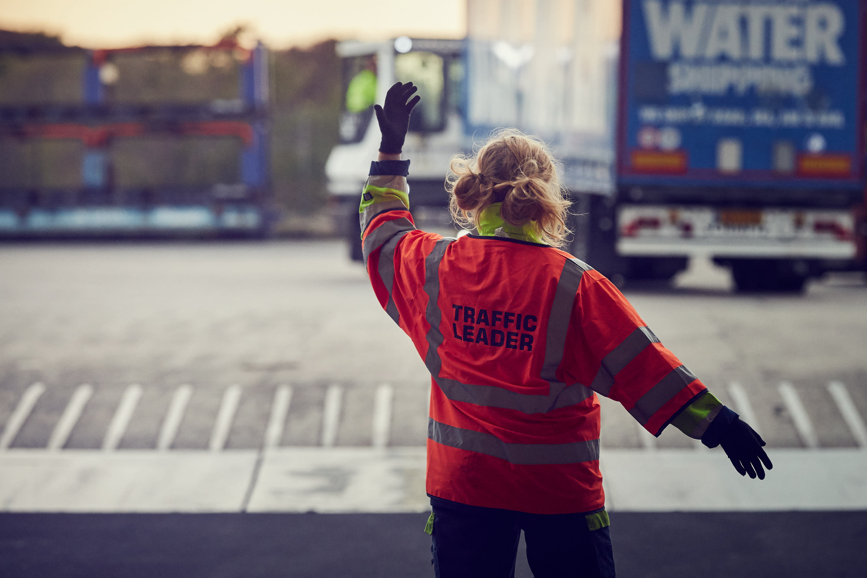 A woman wearing an orange vest that says Traffic Leader signals to a truck in the background DFDS traffic leader